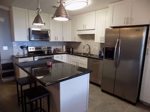A modern kitchen with white cabinets, a stainless steel refrigerator, a microwave, and a large island with stools. - Tubbies on the Bay (Pensacola)