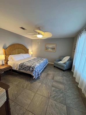 Une chambre d’hôtel avec un lit, une chaise, un ventilateur de plafond, un tableau et une fenêtre avec des rideaux.