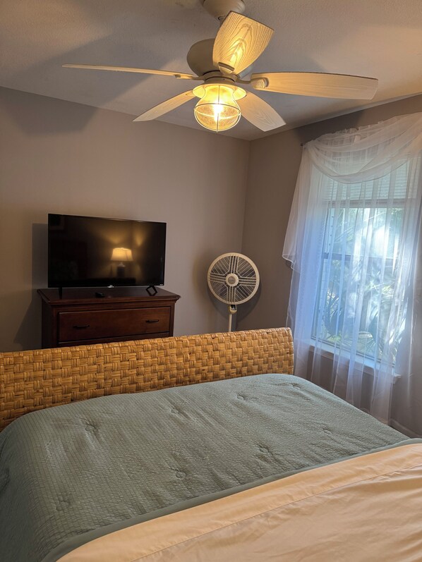 A bedroom with a bed, a television on a dresser, a ceiling fan, and a window with sheer curtains.