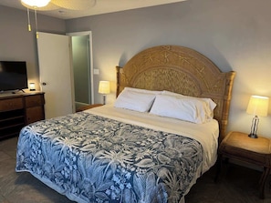 A bedroom with a bed, two bedside lamps, a wooden headboard, a television on a stand, and a door. - Tubbies on the Bay (Pensacola)