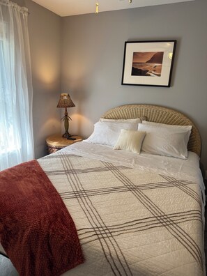 Un dormitorio con cama, lámpara de noche, un cuadro en la pared y una ventana con cortinas.