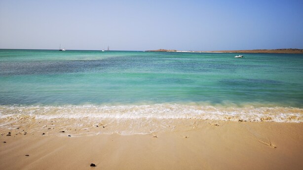 Una playa con agua turquesa cristalina, una orilla de arena y una línea costera distante con vegetación escasa.