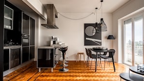 Une cuisine moderne avec un parquet en chevrons, des appareils électroménagers en acier inoxydable et un coin repas offrant une vue dégagée grâce à de grandes fenêtres.