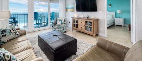 A spacious living room with a large TV, a comfortable sofa set, a wooden cabinet, and a balcony with a view of the sea.