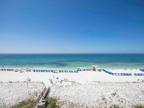 A beach with white sand and clear blue water. There are blue and red umbrellas and lounge chairs along the shore.