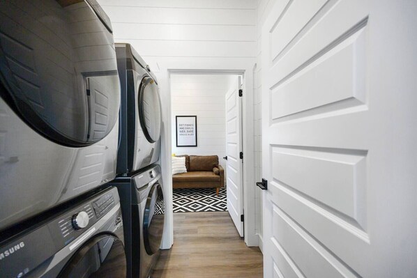 A laundry room with stacked washers and dryers, a patterned rug, and a door leading to a living area with a couch.