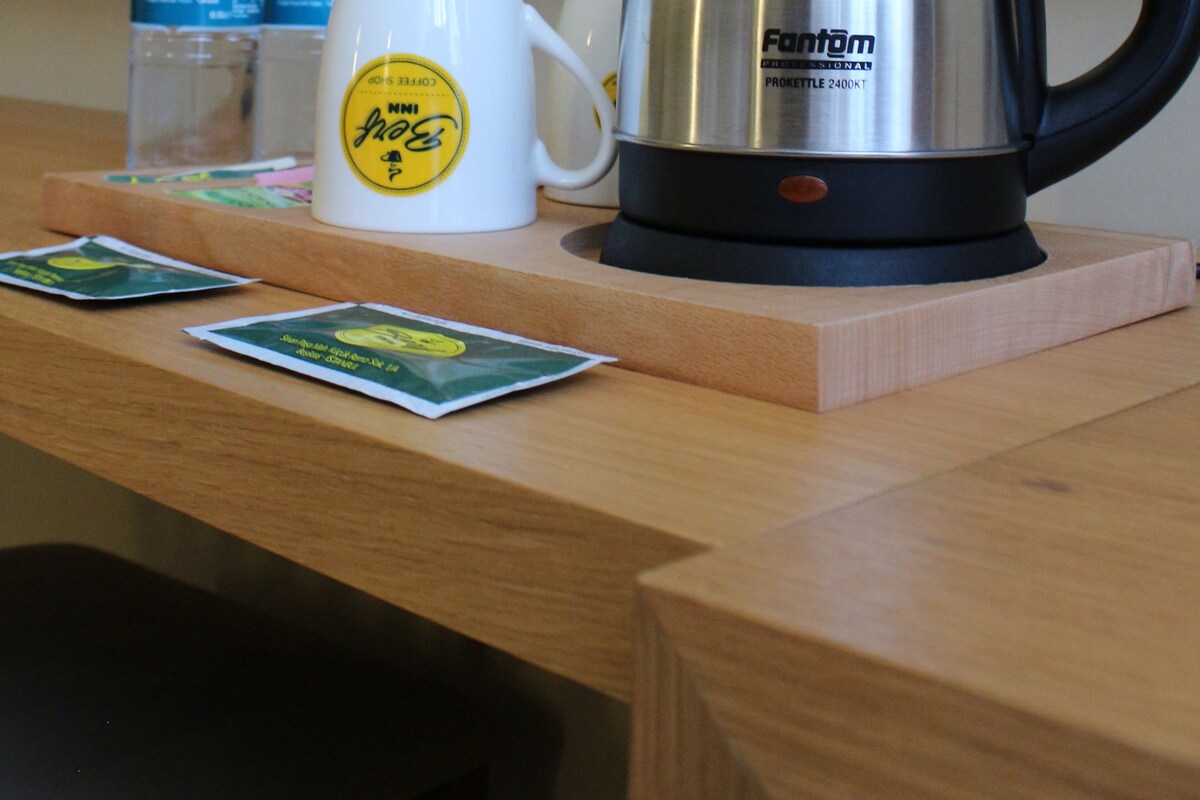 A hotel room with a wooden desk, a stainless steel kettle, two water bottles, and a white mug with a logo.