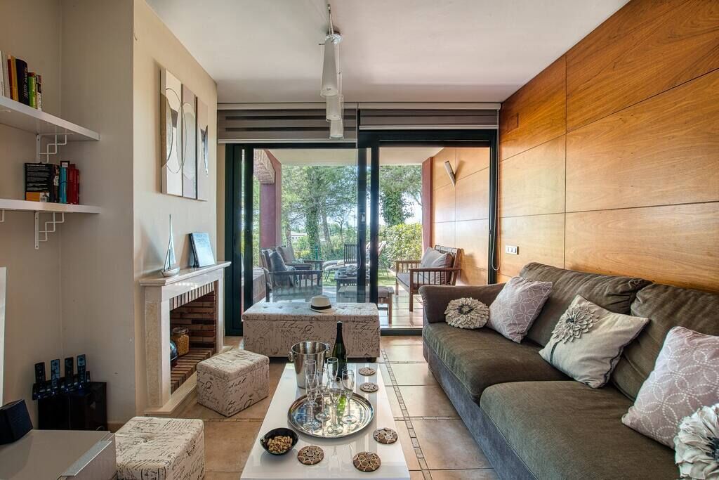 A modern living room with a sofa, coffee table, and fireplace. There are bookshelves and a view of the outdoors.