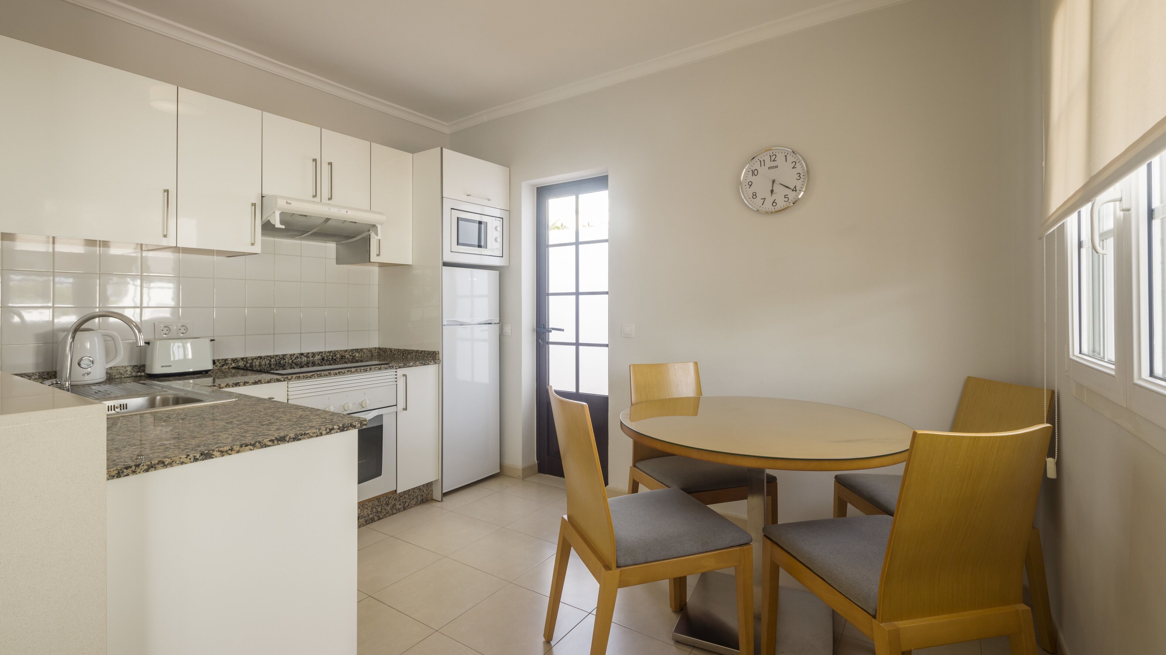 A modern kitchen with white cabinets, a granite countertop, a built-in oven, and a microwave. There is a dining area with a round table and four chairs.