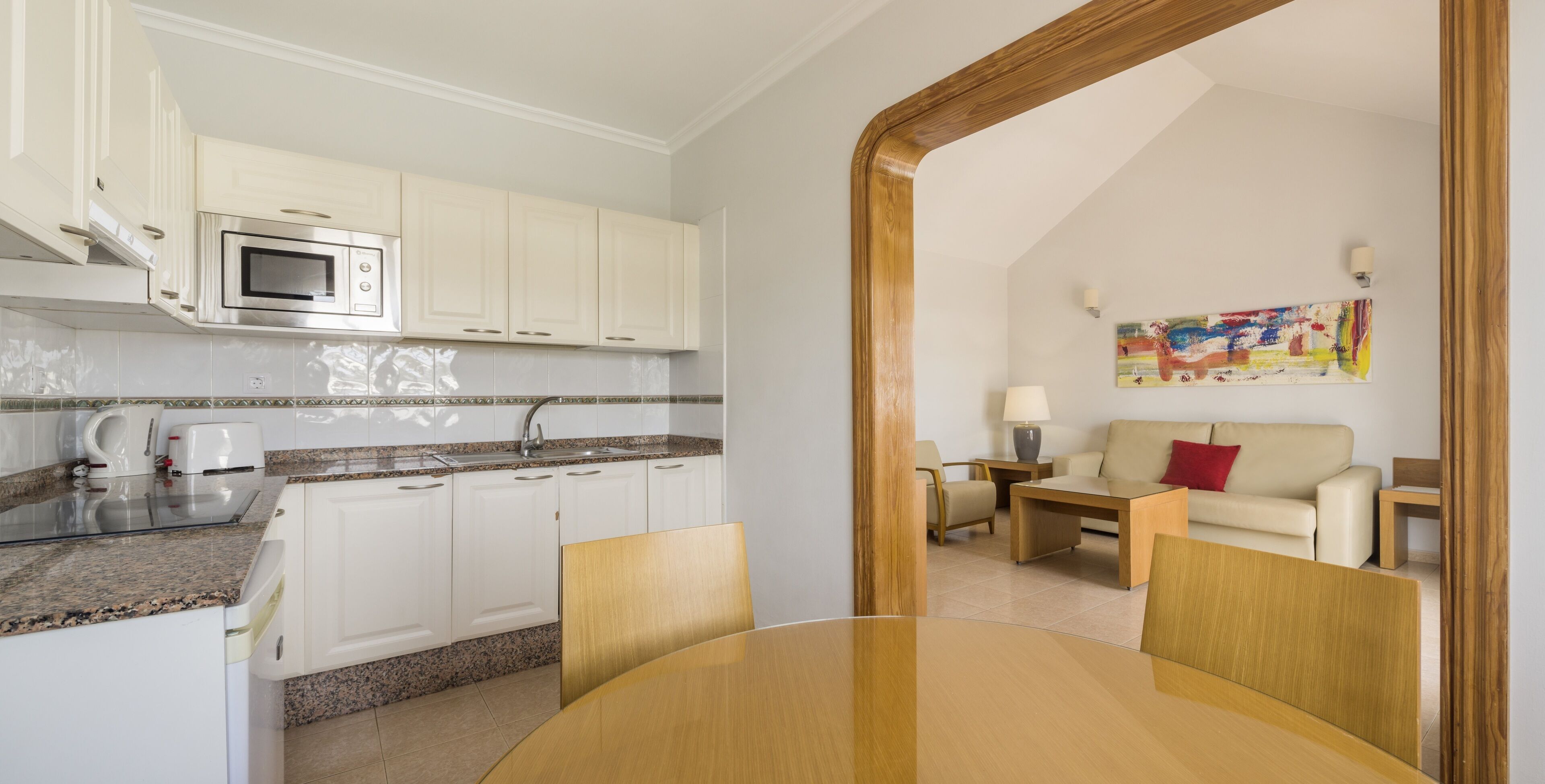 A modern kitchen with white cabinets, a microwave, and a granite countertop. There is a dining area with a round table and chairs, and a beige sofa in the background.