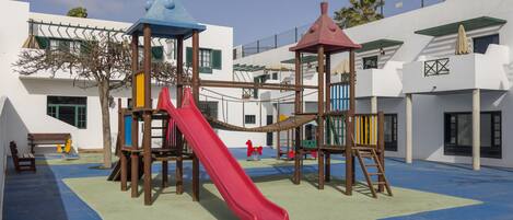 A playground with a red slide and climbing structures, surrounded by white buildings with green shutters and a blue roof.