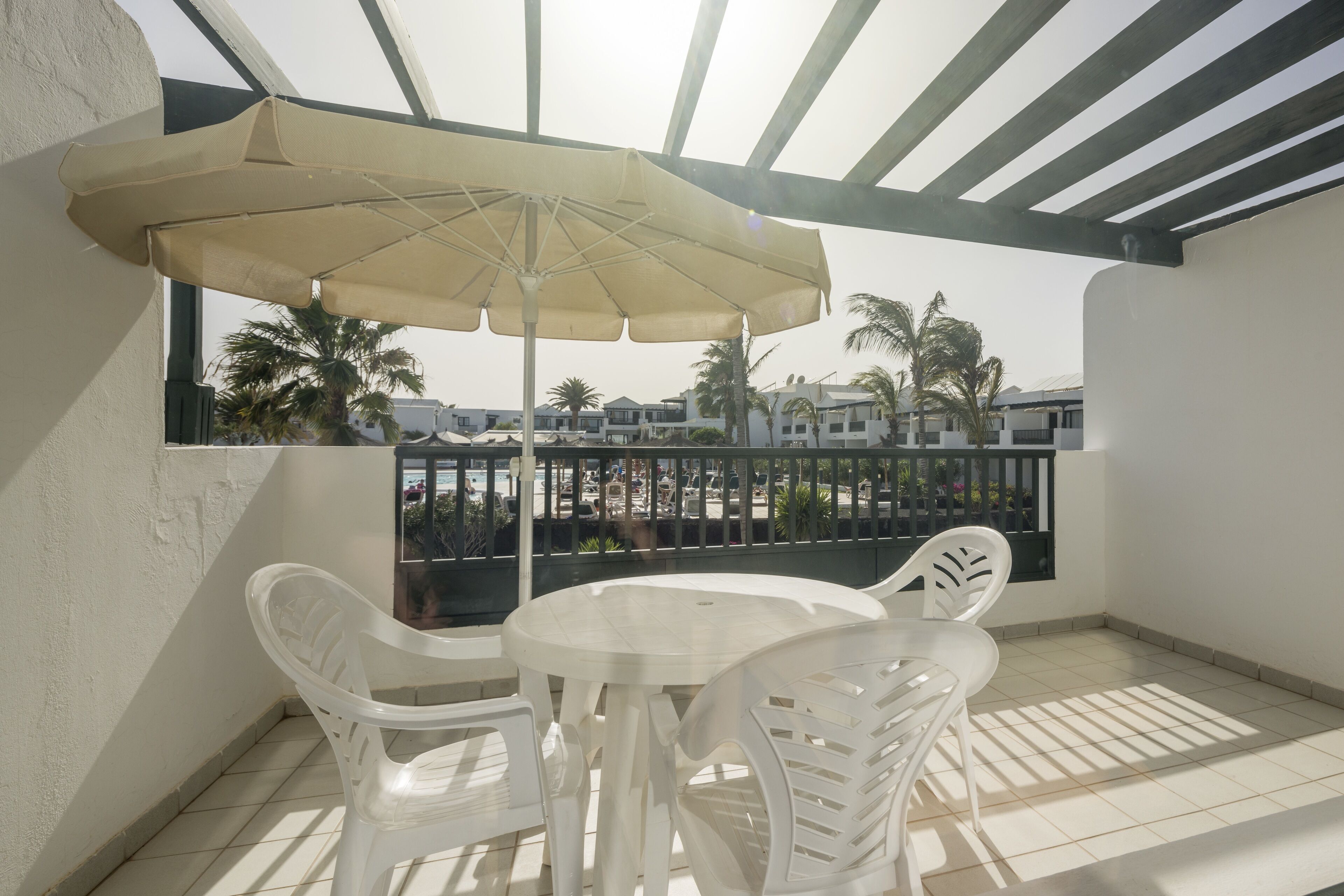 A balcony with a white table and chairs, a beige umbrella, and a view of palm trees and buildings.