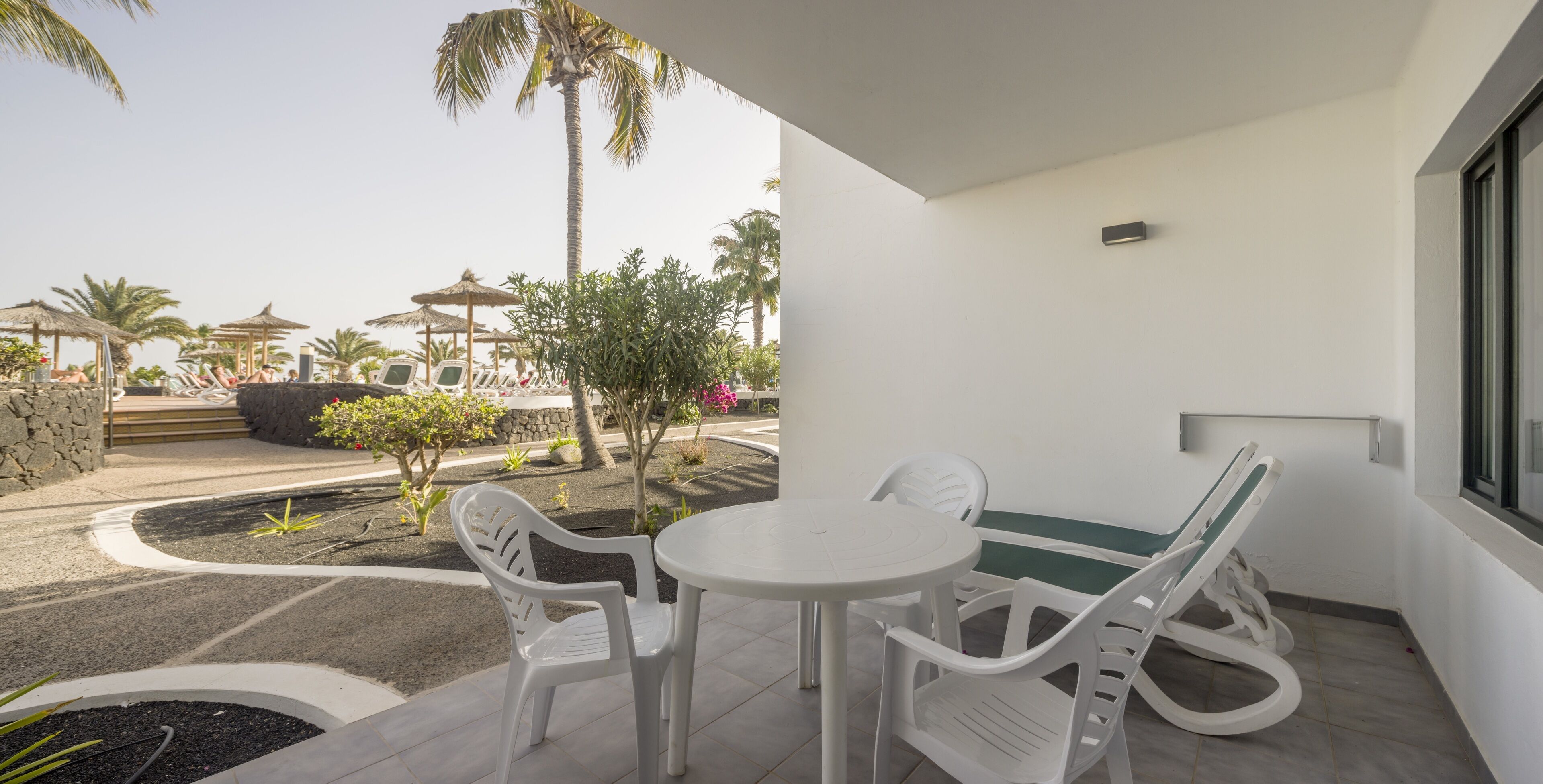 A patio with a table and chairs, a reclining chair, and a view of a pool area with palm trees and a gazebo.