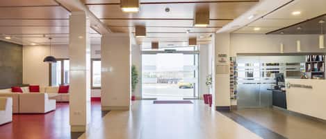 A modern hotel lobby with a reception desk, a seating area with red cushions, and a view of the outside.