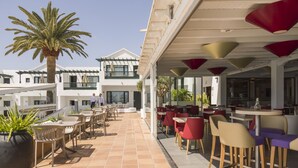 Un patio al aire libre con mesas y sillas, una palmera y un edificio con amplias ventanas y un balcón.