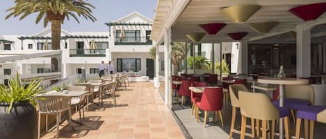 An outdoor patio area with tables and chairs, a palm tree, and a building with large windows and a balcony.