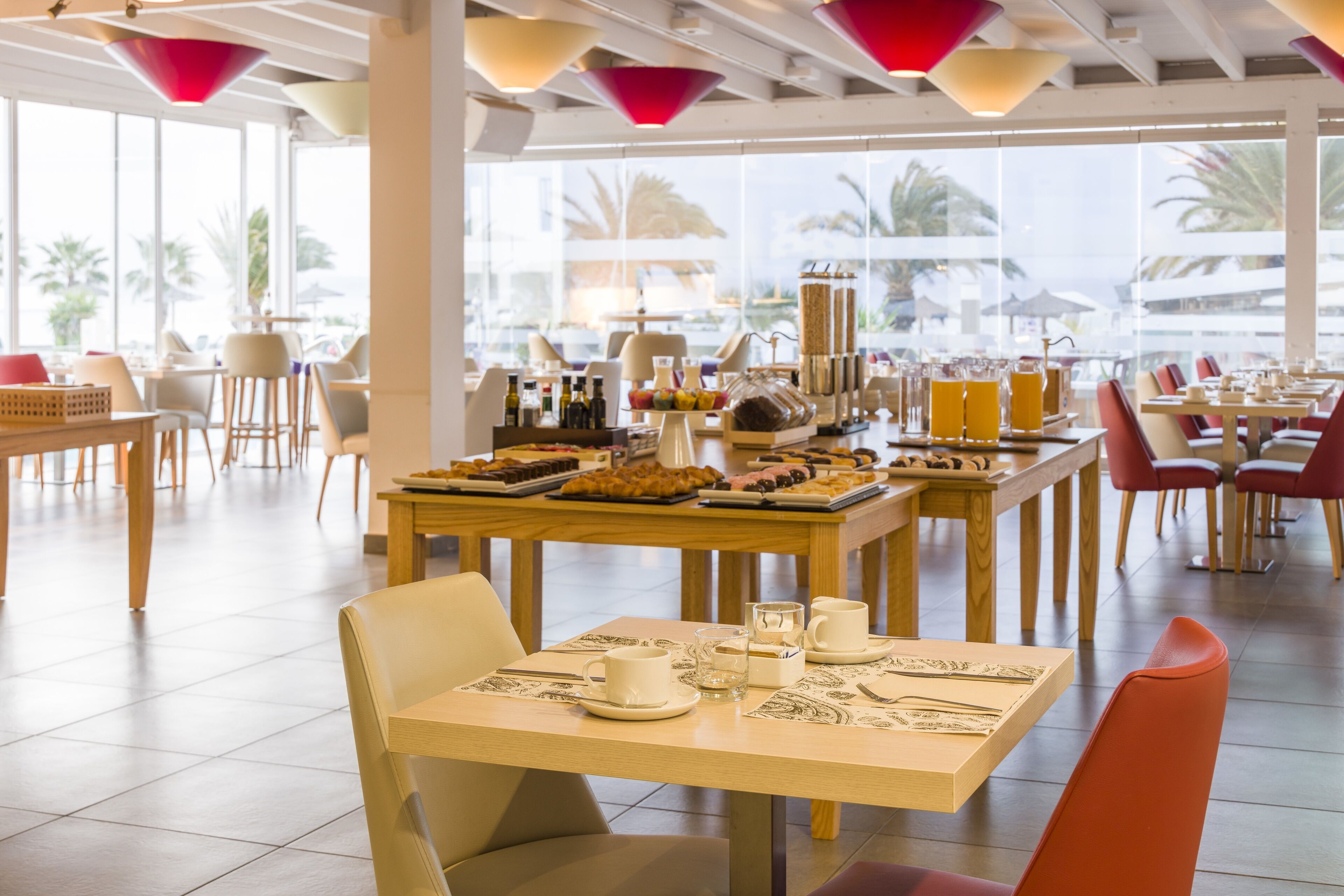 Un comedor amplio con varias mesas y sillas, ventanas grandes que ofrecen vistas a palmeras, y una variedad de opciones de comida y bebidas.