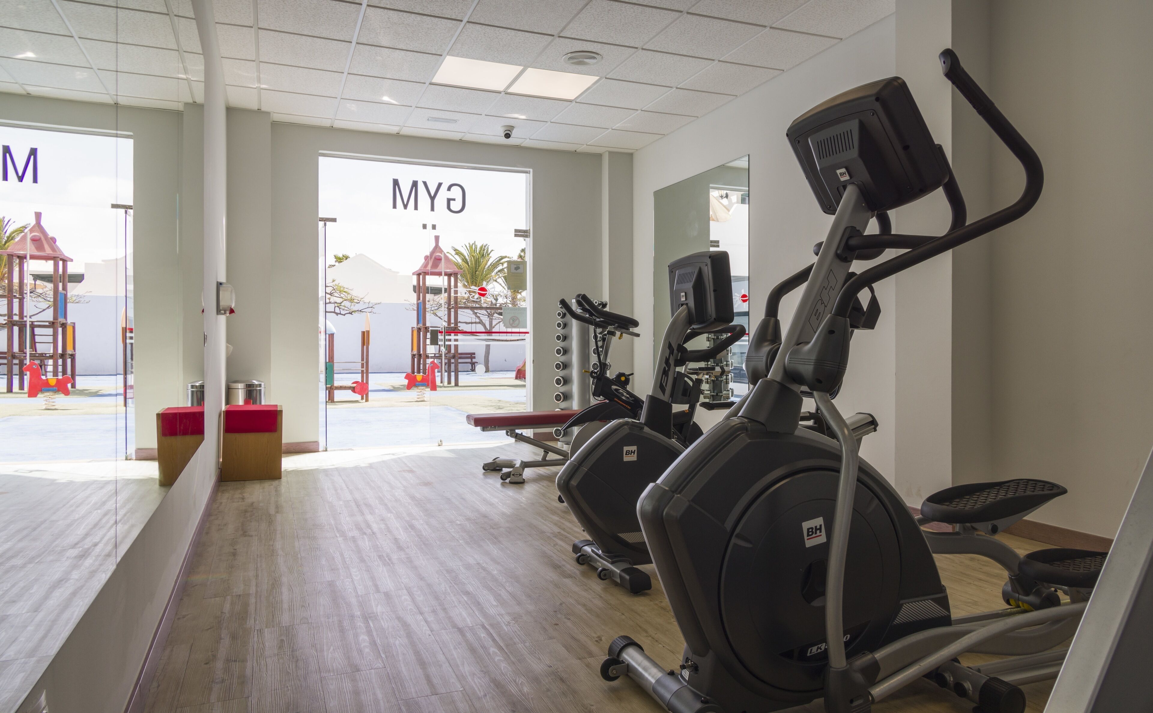 Un gimnasio con equipo de ejercicio, piso de madera y vista a un parque infantil exterior.