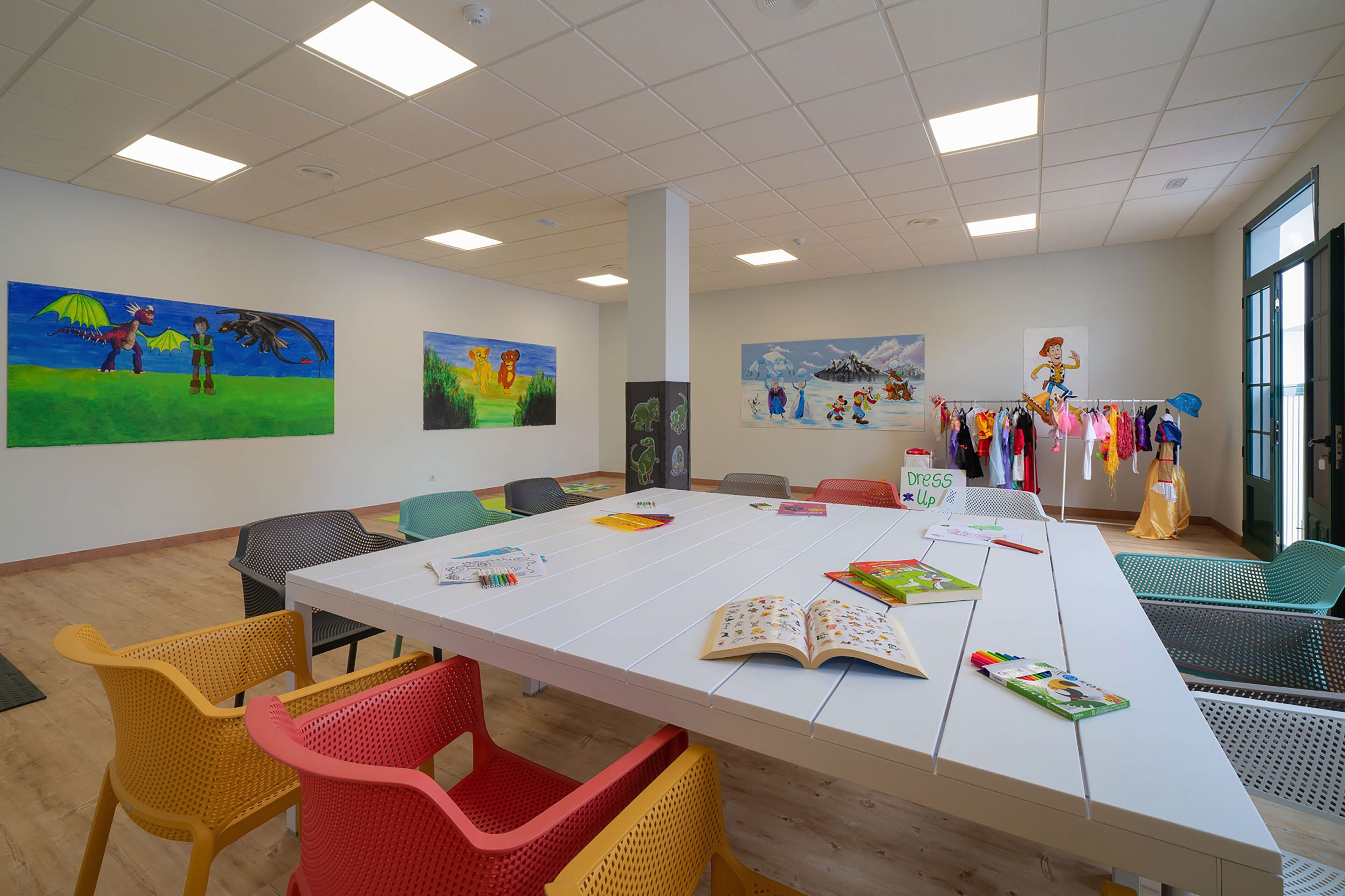 Un aula con una larga mesa, sillas coloridas, carteles educativos y una exposición de ropa infantil.