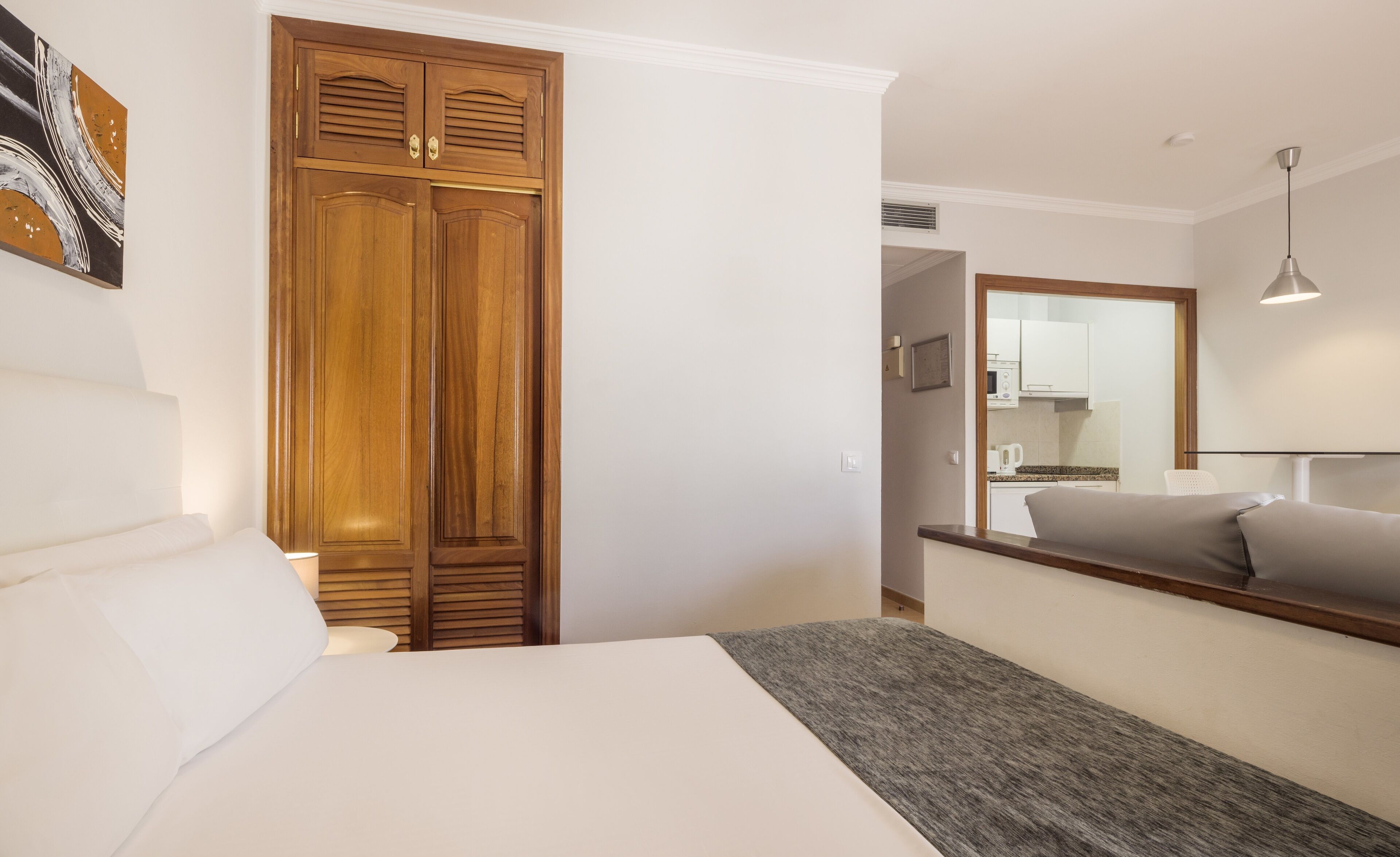 Un dormitorio con cama, armario, mesita de noche, lámpara, sofá y una cocina visible a través de una puerta abierta.