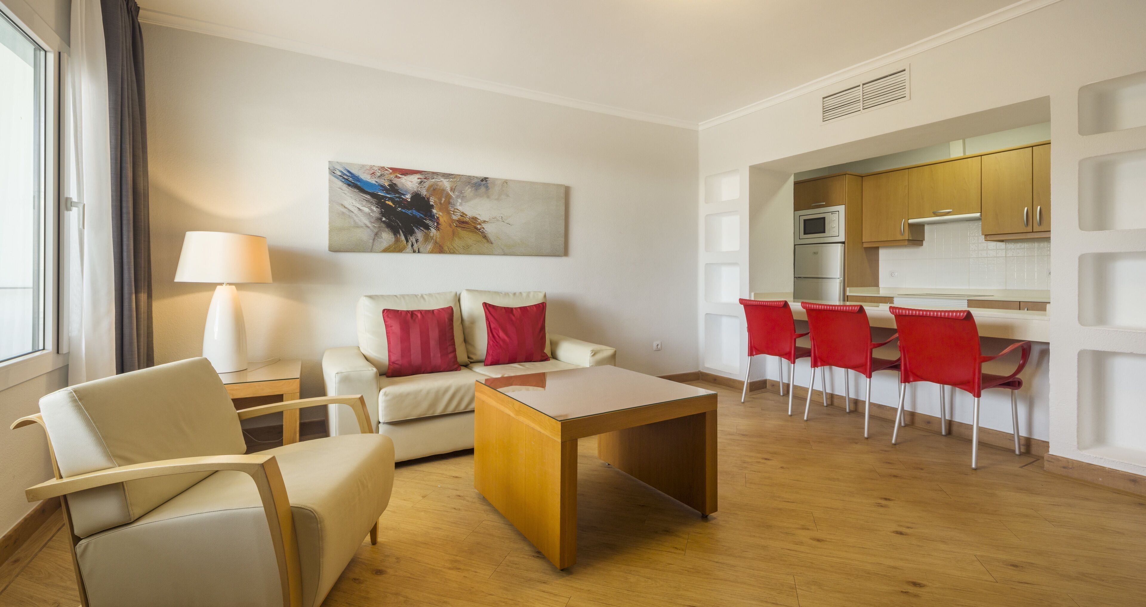 Ein modernes Wohnzimmer mit einer Couch, einem Couchtisch und roten Sesseln. Im Hintergrund ist eine Küche mit Holzschränken und einer Mikrowelle zu sehen.