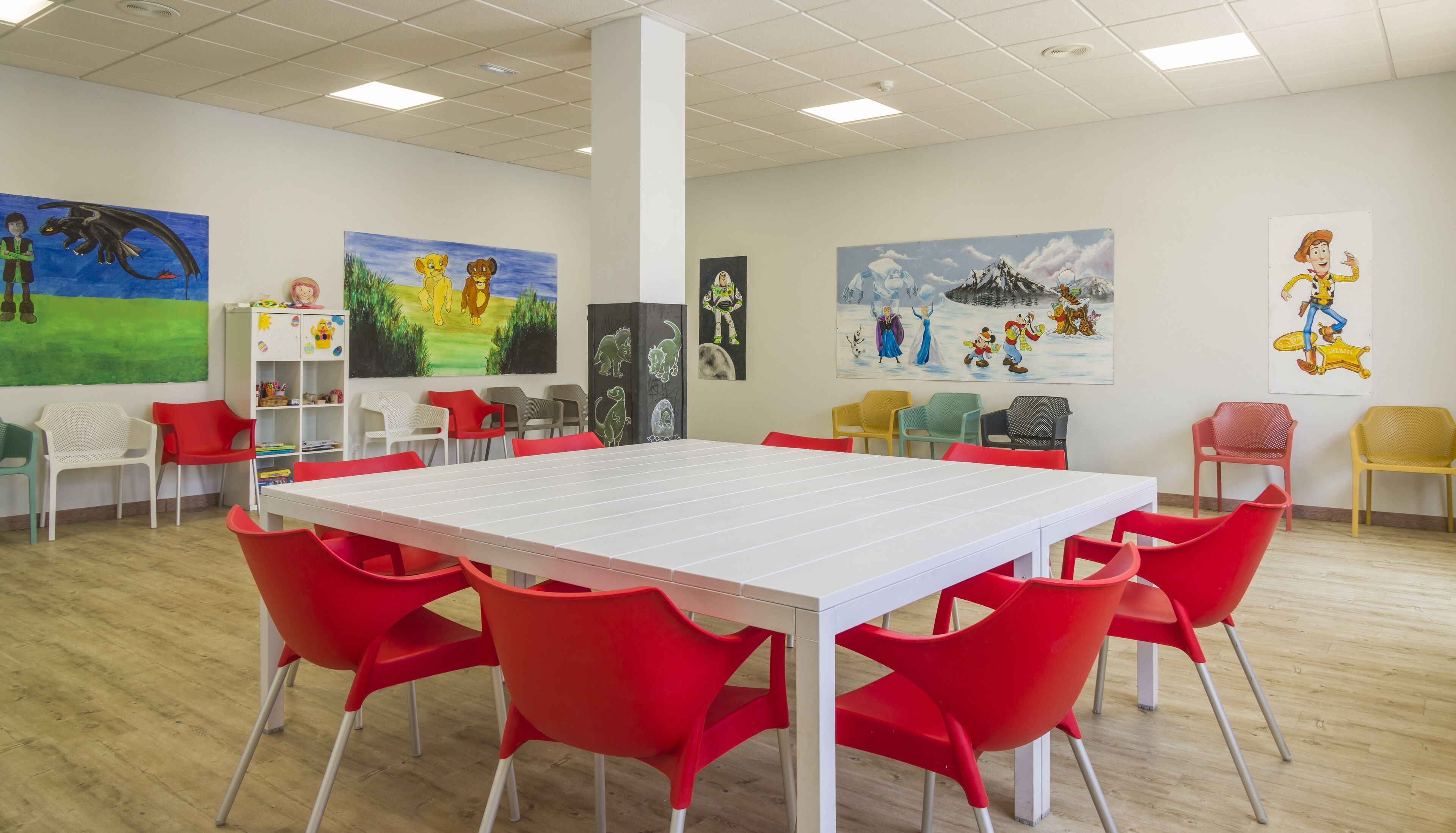 Un aula con una mesa blanca y sillas rojas, rodeada de carteles educativos de colores en las paredes.