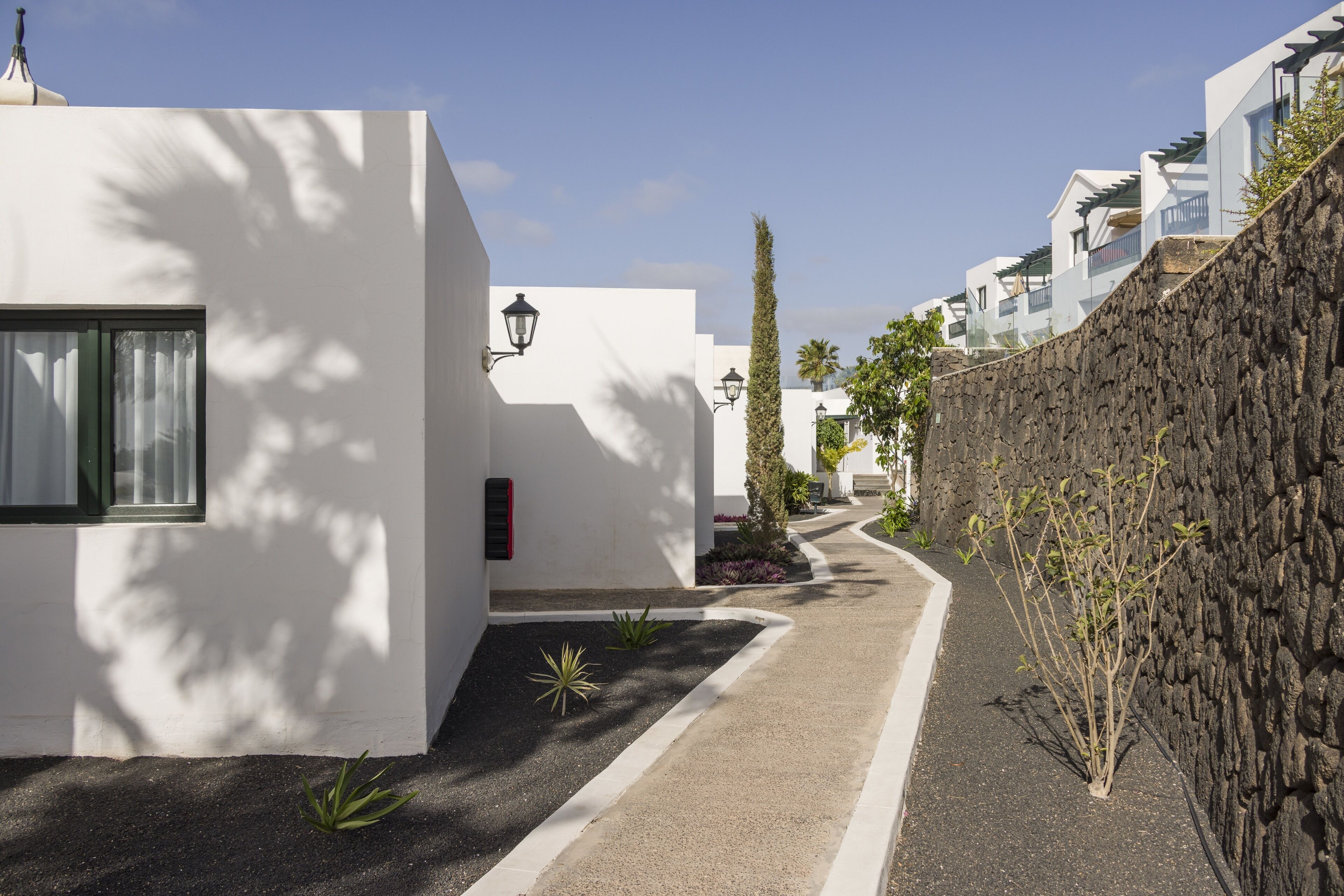 A narrow pathway flanked by white buildings with a stone wall on one side and a row of windows on the other.