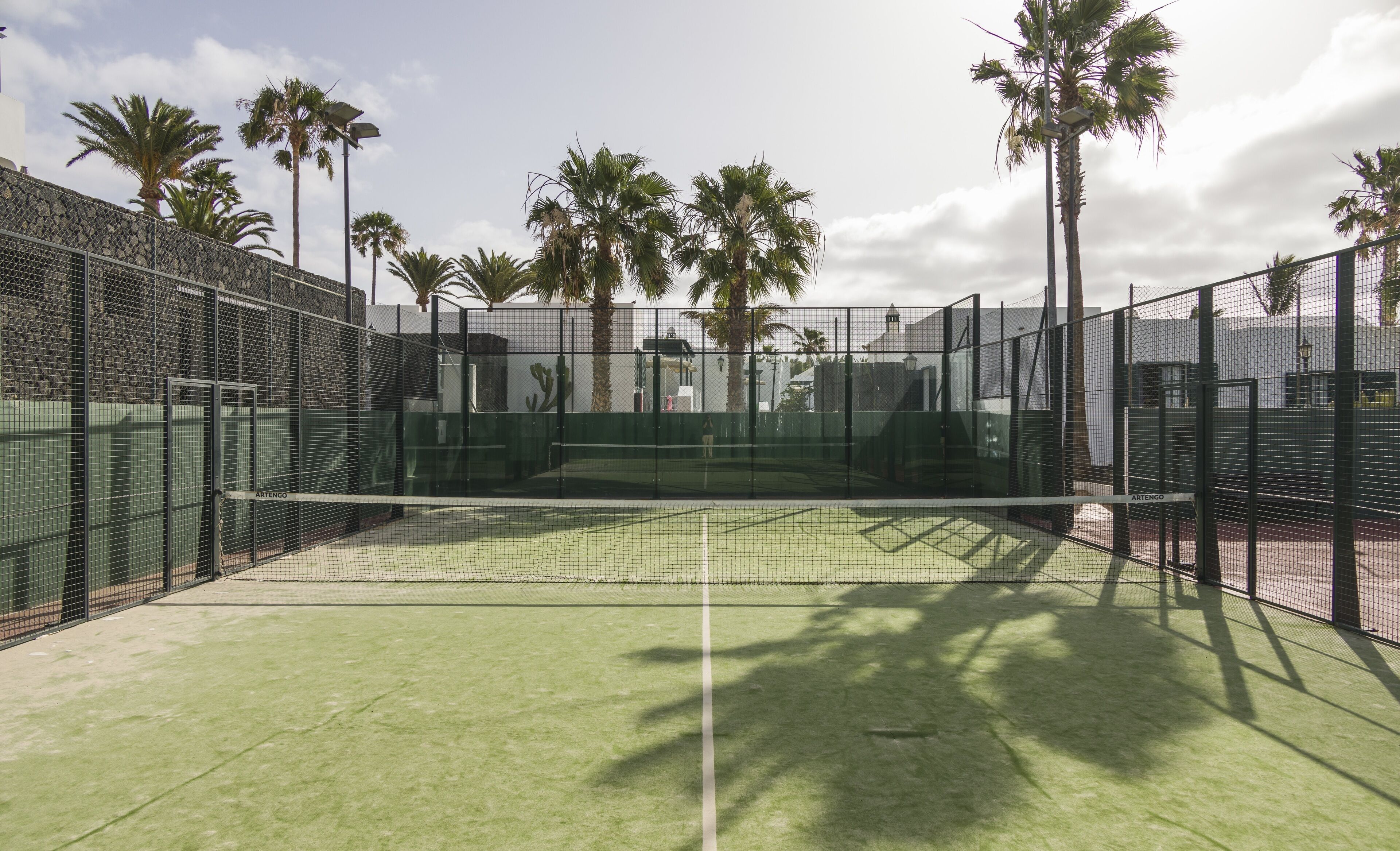 An outdoor tennis court with a net, surrounded by a metal fence and palm trees.