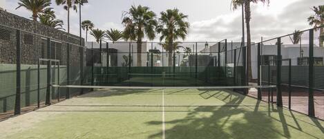 An outdoor tennis court with a net, surrounded by a metal fence and palm trees.