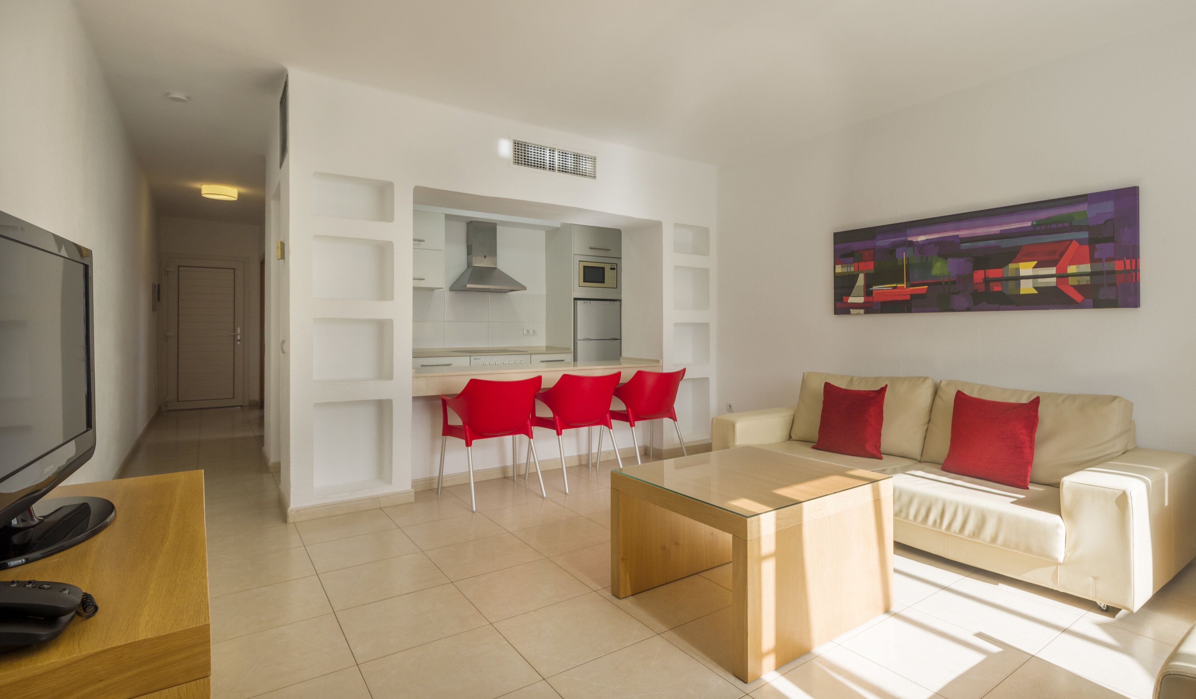 A modern living room with a beige sofa, red chairs, a glass coffee table, and a TV on a wooden table.