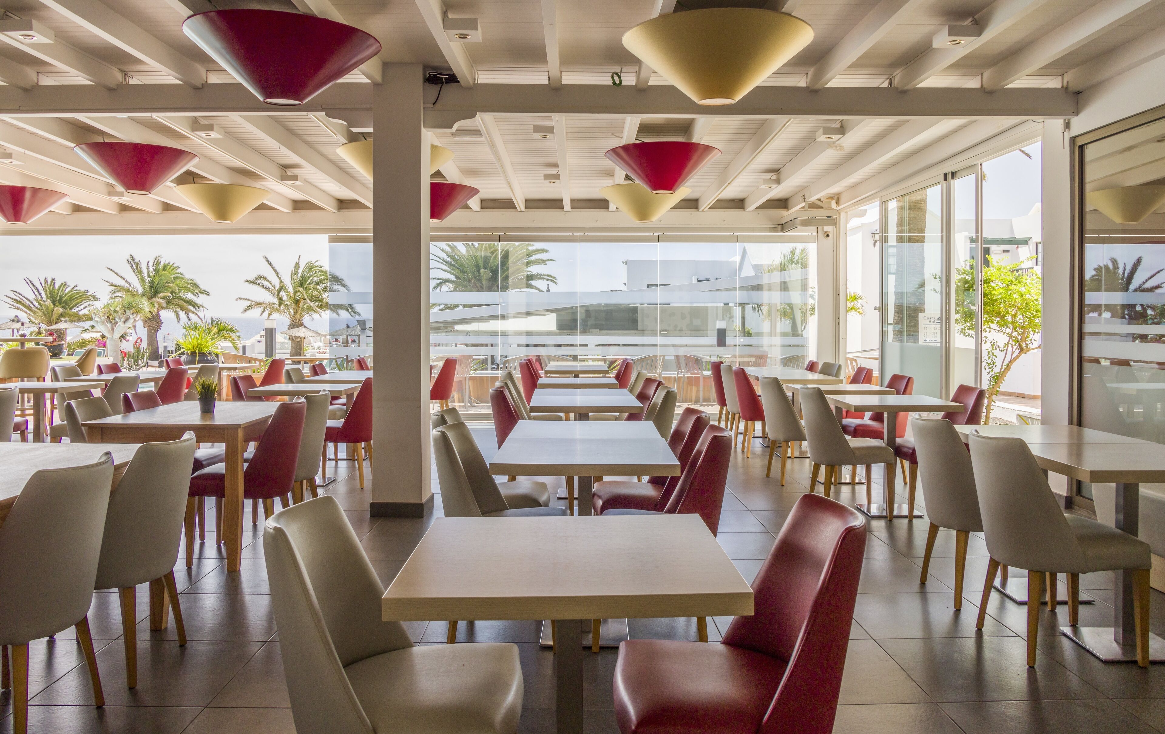 A spacious dining area with multiple tables and chairs, large windows offering a view of palm trees and the sea, and modern pendant lighting.
