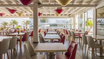 A spacious dining area with multiple tables and chairs, large windows offering a view of palm trees and the sea, and modern pendant lighting.