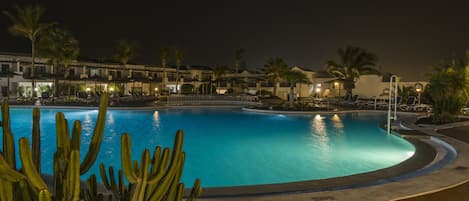 Ein Poolbereich bei Nacht mit klarem, blauem Wasser, umgeben von Palmen und Liegestühlen.