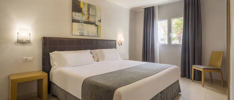 A hotel room with a large bed, a bedside table, a chair, a window with curtains, and a painting on the wall.