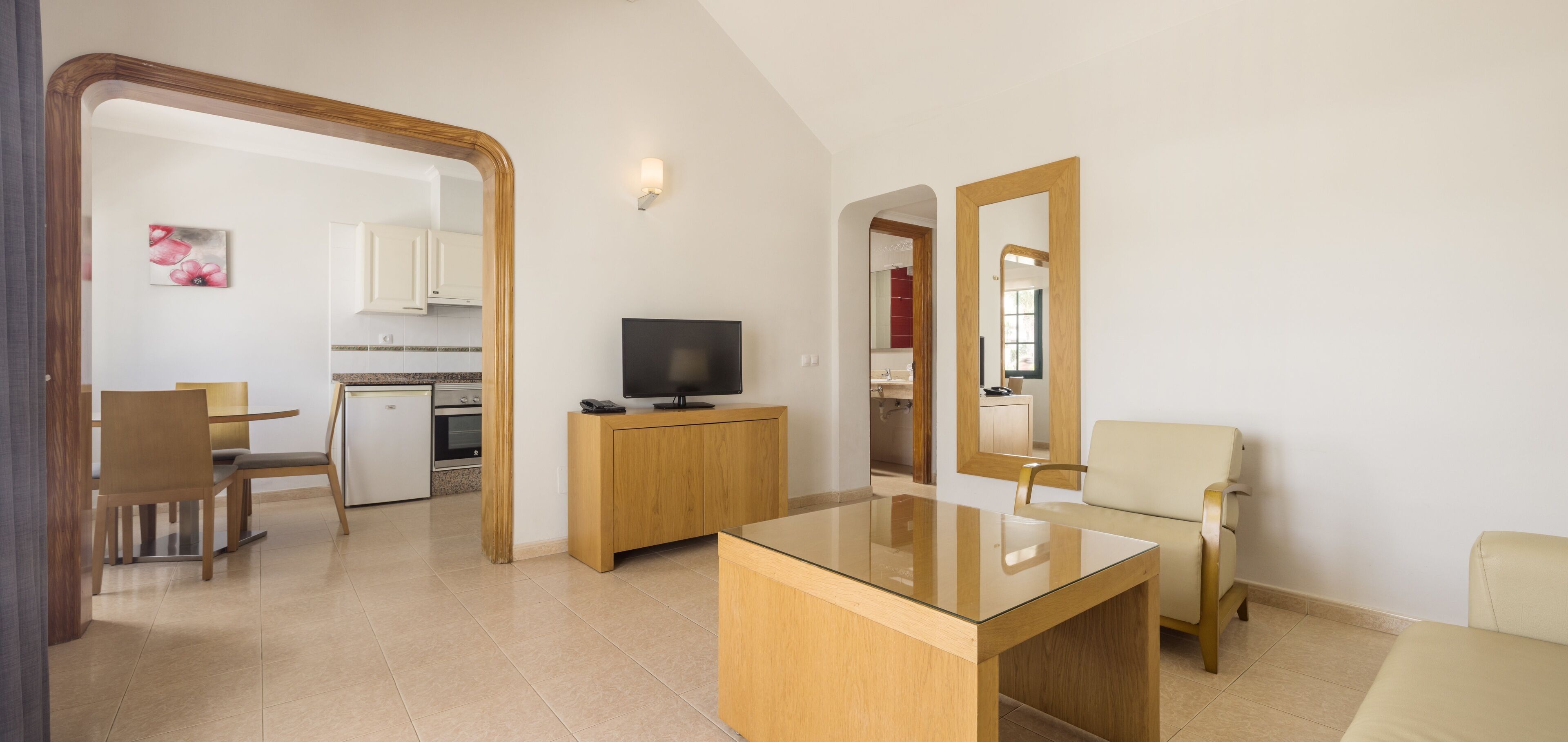 Una sala moderna con una mesa de centro de vidrio, un televisor sobre un mueble de madera, un sofá color beige y un comedor con una mesa y sillas de madera.