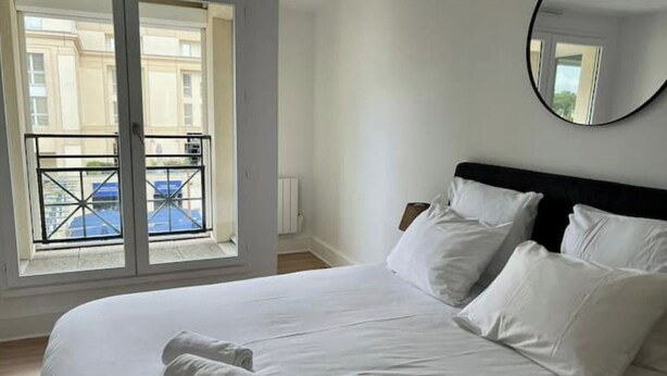 Een hotelkamer met een groot bed, wit beddengoed, een ronde spiegel en een balkon met uitzicht op gebouwen en een blauwe auto.