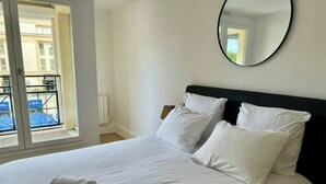 Een net opgemaakt bed met witte lakens en kussens, een ronde spiegel aan de muur, en uitzicht op een balkon met een gebouw op de achtergrond.