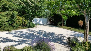 Une allée de gravier mène à un portail blanc, entouré d’une végétation luxuriante et d’arbres.