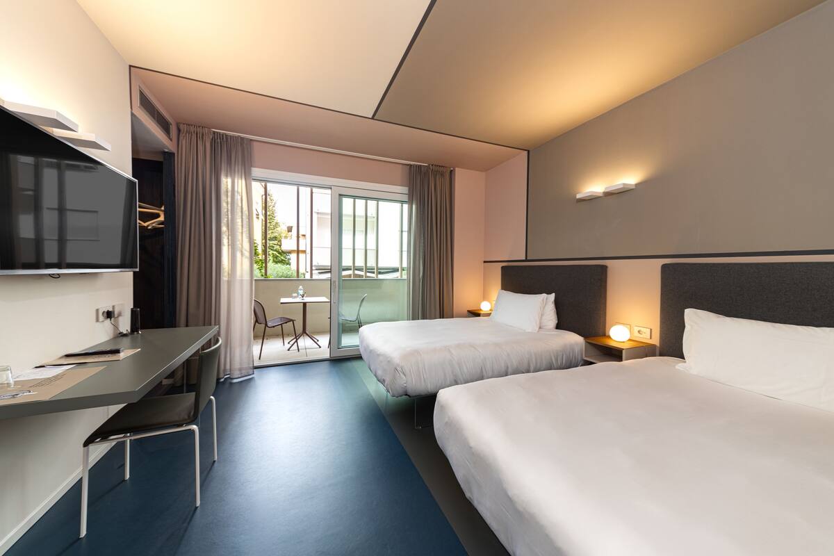 A hotel room with two beds, a desk, a chair, a TV, and a balcony with a view.