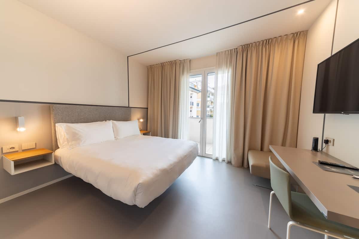 A modern hotel room with a large bed, bedside tables, a desk with a chair, a TV, and a sliding glass door to the outside.