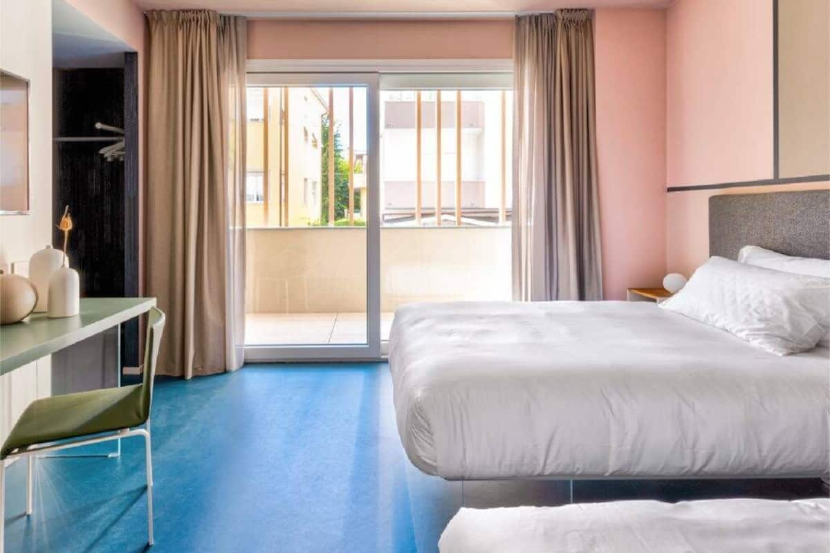 A hotel room with a bed, a desk, a chair, and a balcony with curtains.
