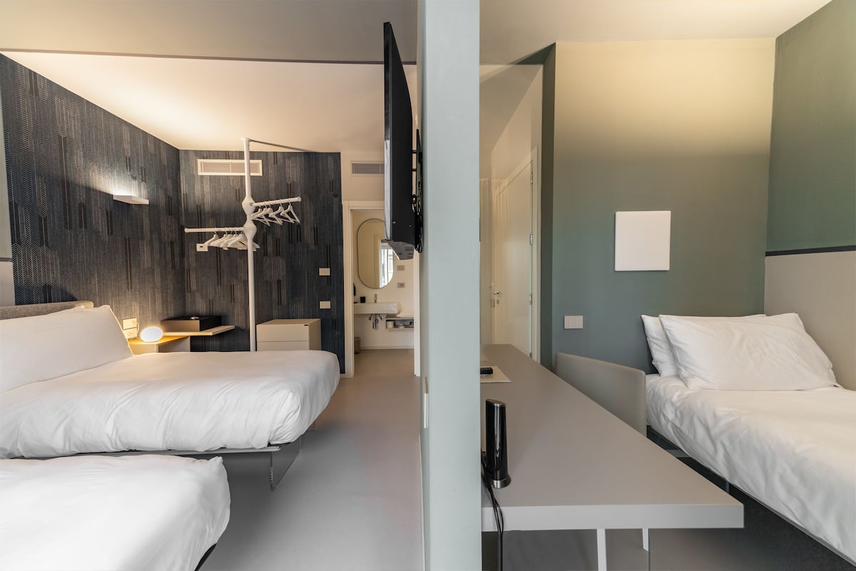 A modern hotel room with two beds, a desk, a mirror, and a bathroom visible through an open door.