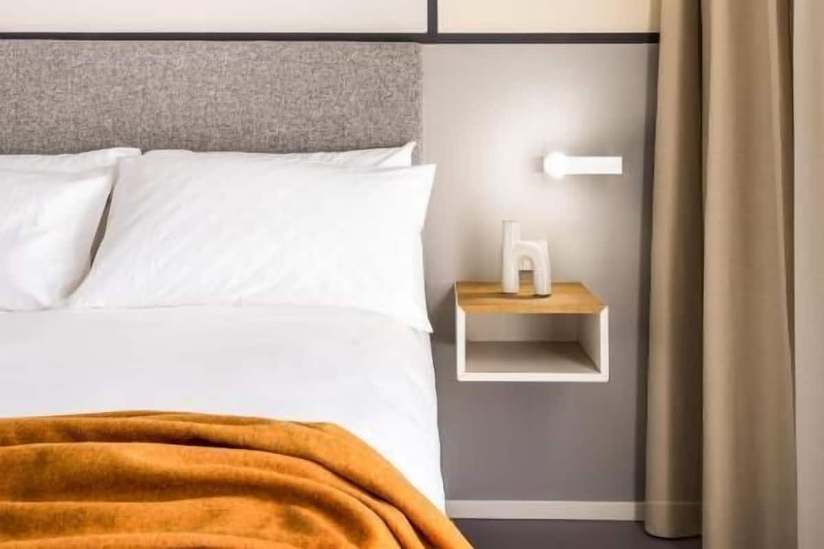 A modern hotel room with a bed, a nightstand, and a wall-mounted light.
