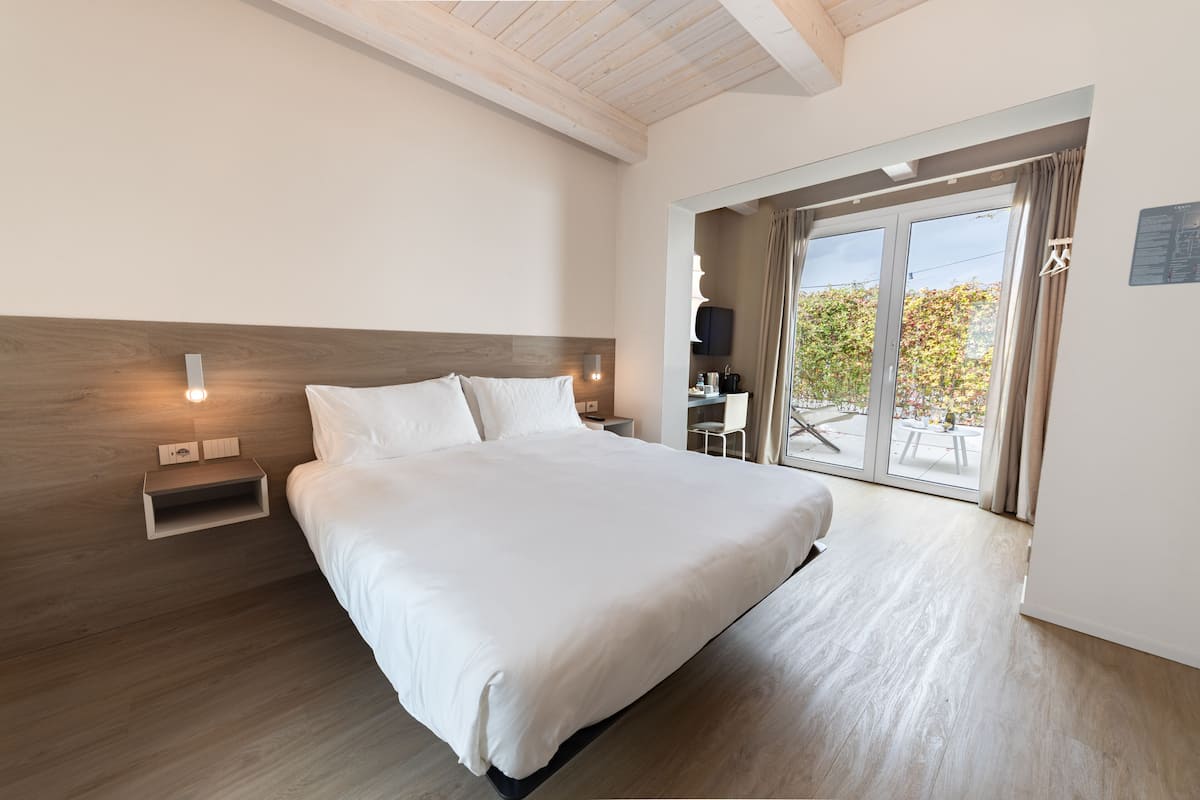 A modern hotel room with a large bed, wooden headboard, bedside tables, and a balcony with a view of trees.