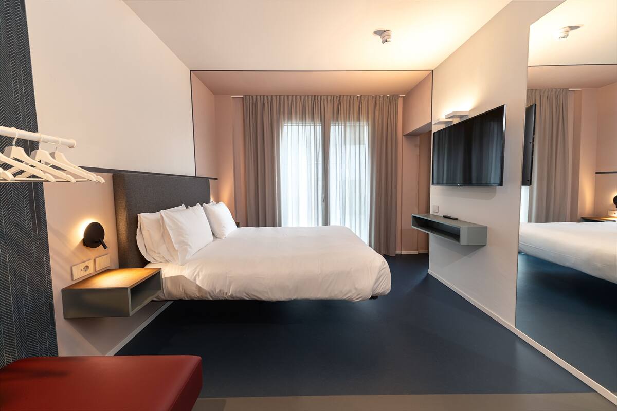 A modern hotel room with a large bed, a bedside table, a TV, and a wardrobe.