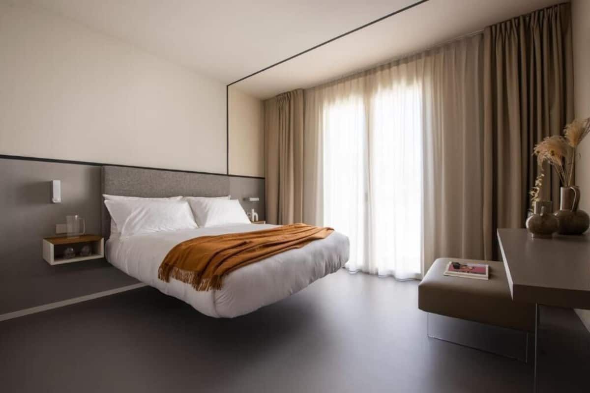A modern hotel room with a large bed, a bedside table, a desk, and a window with curtains.