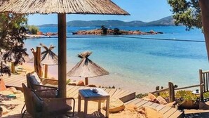 Eine Holzterrasse mit Reetsonnenuren, Liegestühlen und Blick auf ein ruhiges Meer und entfernte Inseln.