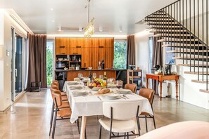 Uma área de jantar moderna com uma mesa comprida posta para uma refeição, uma cozinha ao fundo e uma escada com corrimão.