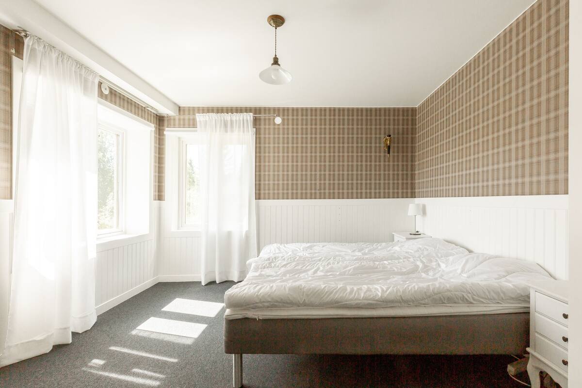 Un dormitorio con una cama, una mesita de noche, una lámpara y un ventanal con cortinas.