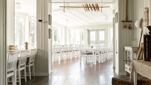 A spacious room with white wooden walls, a high ceiling, and large windows allowing natural light. There are multiple white tables and chairs arranged for dining or meetings. - Edsåsgården (Undersaker)